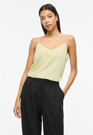 Vrouw met lang donker haar, gekleed in een lichtgele mouwloze top en hooggetailleerde zwarte broek, staand met één hand in haar zak.