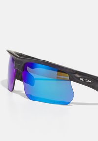 Schwarze Sportsonnenbrille mit großen, gebogenen blauen Gläsern und lila Farbverlauf. Leichtes Design mit strukturierten Ohrenpolstern und markenspezifischen Details.