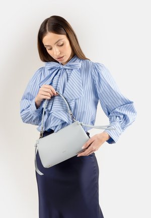 Donna con blusa a righe blu e gonna scura che tiene e guarda una piccola borsa grigio chiaro su uno sfondo neutro.