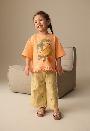 Fiatal lány mosolyog, narancssárga banánmintás pólót, csíkos gyümölcsmintás nadrágot és rózsaarany szandált visel, bézs szék mellett áll.