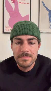Dunkler Pullover, grüne Strickmütze, helle Haut, kurze Haare und ein Stoppelschnurrbart. Der Hintergrund zeigt gerahmte Poster mit Text und Illustrationen.