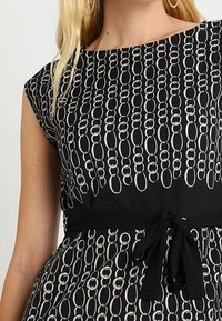 Kobieta w czarnej sukience z białymi, nakładającymi się owalnymi wzorami, z rękawami na kapelusz, czarnym paskiem w talii i złotymi kolczykami w kształcie kół.