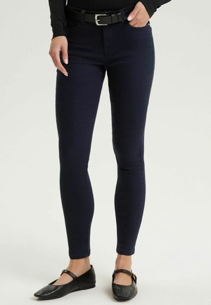 Kobieta ubrana w dopasowane ciemnogranatowe spodnie, czarną bluzkę z długim rękawem, czarny pasek oraz czarne baleriny typu Mary Jane stoi na tle jednolitego tła.