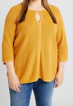 Mosterdgele gebreide top met 3/4 mouwen, voorzien van een keyhole halslijn en een relaxte pasvorm, gecombineerd met blauwe jeans voor contrast.