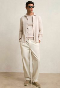 Cardigan bej deschis cu detalii de butoane peste un pulover asortat, combinat cu pantaloni albi și pantofi slip-on gri. Ochelari de soare negri completează ținuta.