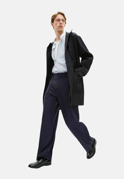 Manteau noir imperméable avec capuche, chemise rayée bleu clair, pantalon bleu marine et chaussures de ville noires ; mannequin en position de marche sur fond blanc.