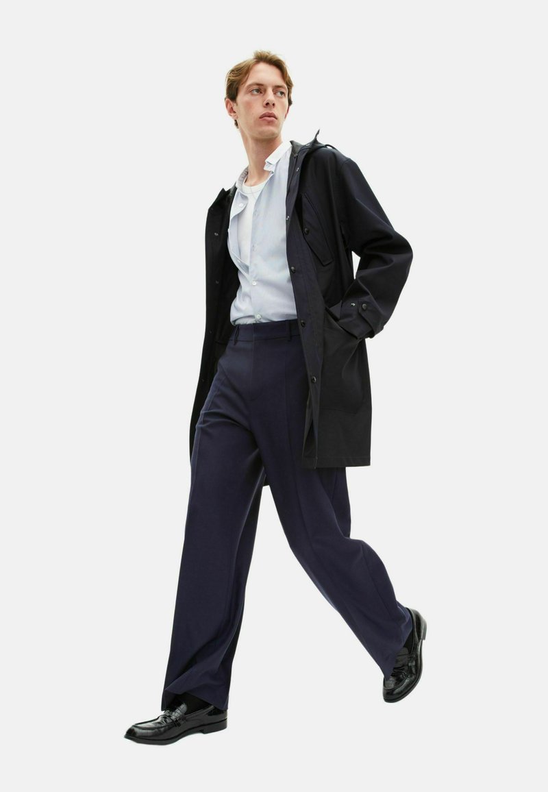 Abrigo negro impermeable con capucha, camisa a rayas azul claro, pantalones navy y zapatos de vestir negros; modelo en una postura de caminata frente a un fondo blanco.