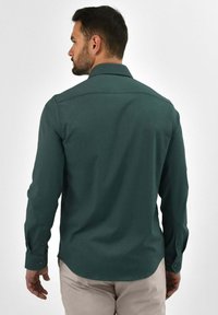 Donkergroen overhemduits met lange mouwen, voorzien van een button-down kraag, een gladde textuur en een getailleerde pasvorm. Zichtbare stitching en met knopen verstelbare manchetten.