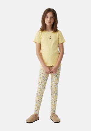 Fille debout, les mains jointes, portant un haut jaune à manches courtes avec un détail fleur, des leggings à motif floral et des chaussures beiges sans lacets.