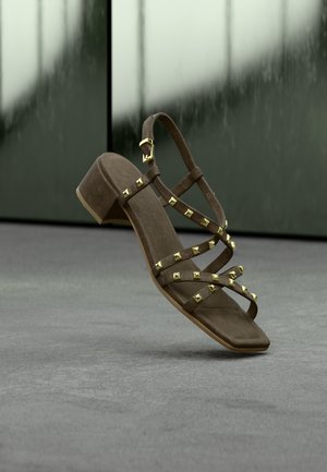 Brūnas zamšādas sandales ar papēdi un zelta piramīdas formas kniedēm, ar krustveida siksnām un potītes sprādzi. Plakanas zolītes un kvadrātveida pirksta dizains.