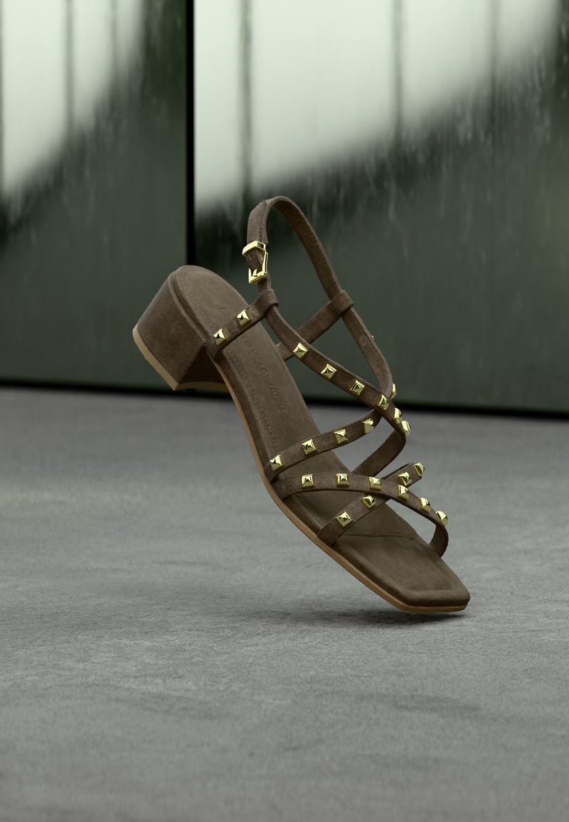Καφέ σουέντ πέδιλο με τακούνι και χρυσά πυραμοειδή τρουκς, με διασταυρούμενους ιμάντες και αγκράφα στον αστράγαλο. Επίπεδη σόλα και τετράγωνο σχήμα δακτύλων.