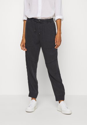 Personne portant un pantalon noir à cordon avec des poignets élastiques et des baskets blanches, associé à une chemise blanche à manches longues retroussées.
