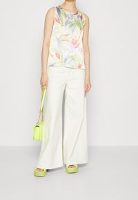 Kobieta ubrana w bezrękawnik w pastelowy wzór liści, białe szerokie spodnie, limonkowe sandały na platformie oraz trzymająca dopasowaną limonkową torebkę.
