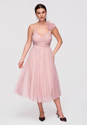 Femme portant une robe midi rose sans manches à pois, avec un détail volanté à l'épaule et des talons noirs pointus, posant une main sur la hanche.