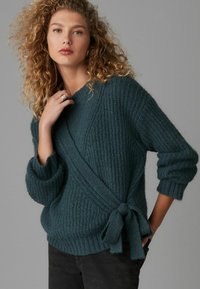Türkisfarbener gestrickter Pullover mit Wickeldesign, strukturiertem Muster und seitlichem Bindeband. Lange Ärmel mit gerippten Bündchen, kombiniert mit dunklen Hosen.