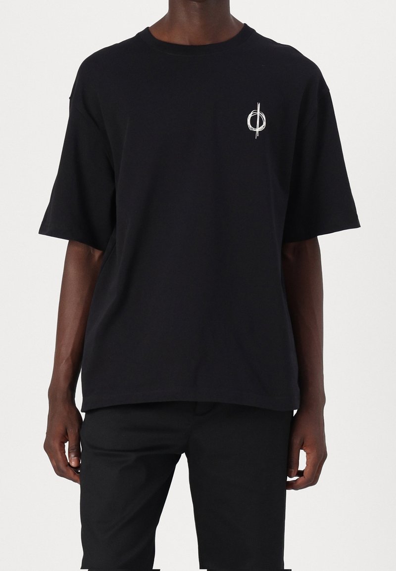 T-shirt noir à manches courtes avec un petit dessin circulaire blanc sur le côté gauche de la poitrine, porté par une personne debout sur un fond uni.