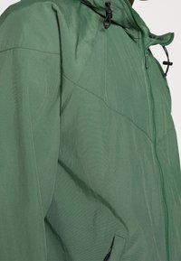 Grüne Kapuzenjacke mit glatter Textur, ausgestattet mit einem Reißverschluss auf der Vorderseite und Kordelzügen an der Kapuze. Nähte sind auf der Brust sichtbar.