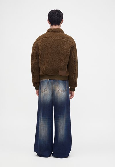 Personne debout de dos, portant une veste marron texturée et un jean bleu délavé à jambes larges sur fond blanc.