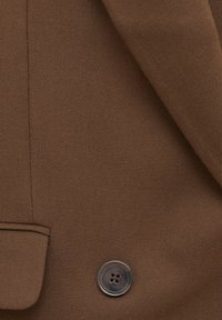 Gros plan d'une veste en tissu marron avec un bouton foncé à quatre trous et un rabat de poche cousu.
