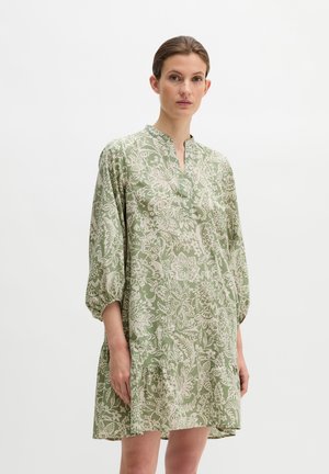 Frau in einem hellgrünen Kleid mit Blumenmuster, dreiviertellangen, gepufften Ärmeln und kleinem V-Ausschnitt, stehend vor einem schlichten weißen Hintergrund.