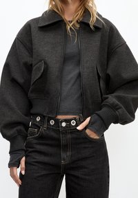 Veste bombardier grise raccourcie avec une finition texturée, dotée d'une fermeture éclair frontale, de grandes poches et de poignets côtelés, associée à un jean noir taille haute.