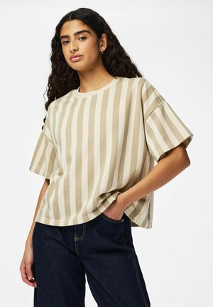 Jeune femme aux cheveux longs et bouclés, portant un T-shirt rayé beige et blanc à rayures verticales et un jean bleu foncé, posant avec une main dans la poche.