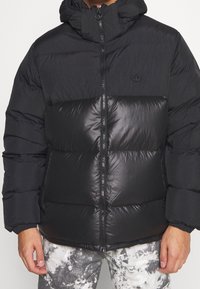 Svart puffjacka med dragkedja, med en texturerad överdel och slät nederdel, utrustad med huva och elastiska ärmslut.