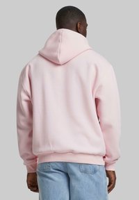 Lys rosa hettegenser med stor hette, ribbestrikkede mansjetter og kant. Myk tekstur og avslappet passform. Brukt over lyse blå jeans.