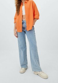 Vrouw in een losse oranje button-up shirt over een witte crop top, lichte blauwe wijduitlopende jeans en crèmekleurige chunky loafers, staand op een witte vloer.