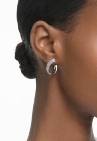 Silver hoopörhänge med en böjd design, som har en rad klara ädelstenar längs framkanten, mot en kortklippt frisyr.