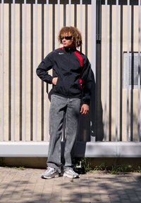 Veste coupe-vent noire avec des accents rouges, jeans gris à jambe droite, et baskets blanches. Modèle se tient devant un mur rayé, portant des lunettes de soleil.