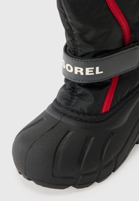 Bota impermeable negra con acentos rojos, que presenta una suela de goma texturizada y una correa gris con marca. Parte superior acolchada para aislamiento.