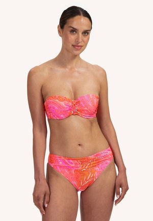 Kobieta w bezramkowym różowo-pomarańczowym bikini w stylu paisley, stojąca z delikatnym uśmiechem i rękami luźno opuszczonymi wzdłuż ciała.