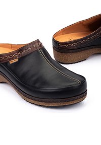 Sabots en cuir noir avec une bande en cuir marron cousue et une semelle semblable au liège. Présente des coutures visibles et une semelle texturée.