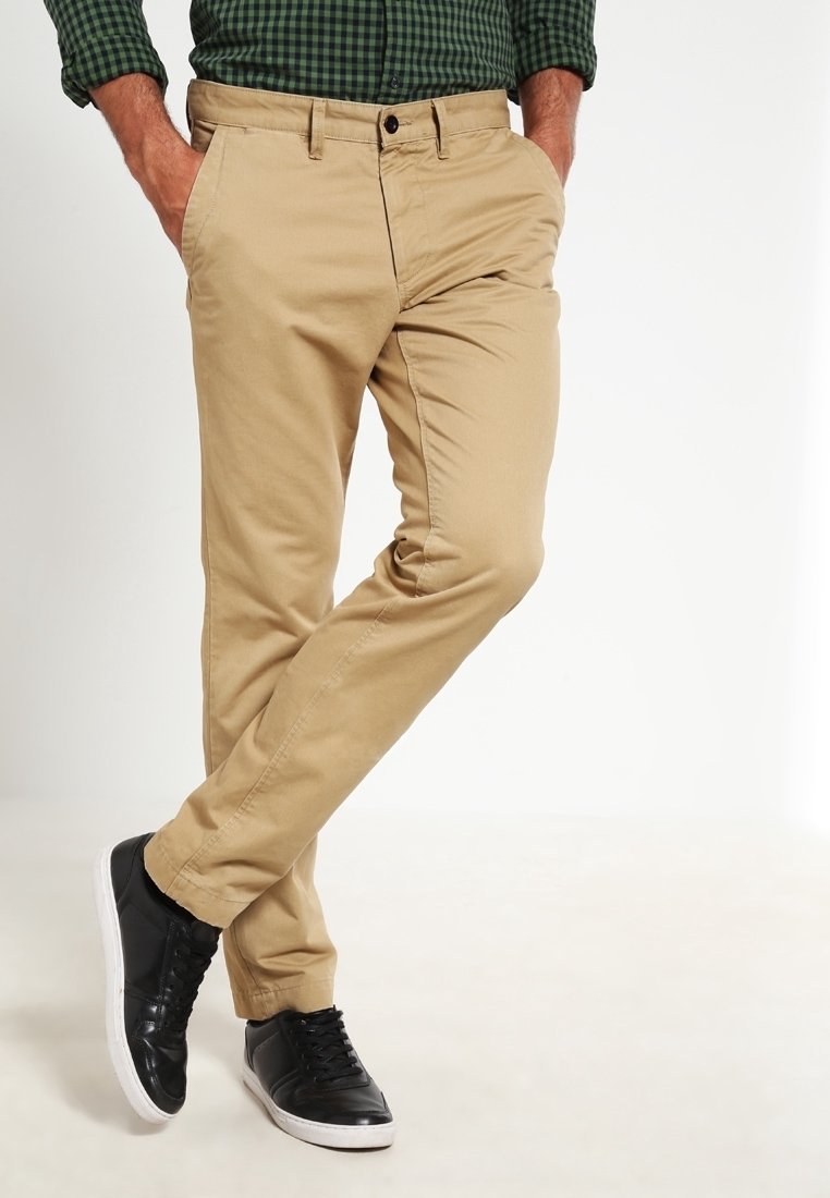 Khaki chino kalhoty s úzkým střihem, s bočními kapsami, zapínáním na knoflík a v kombinaci s černými teniskami a zelenou kostkovanou košilí.