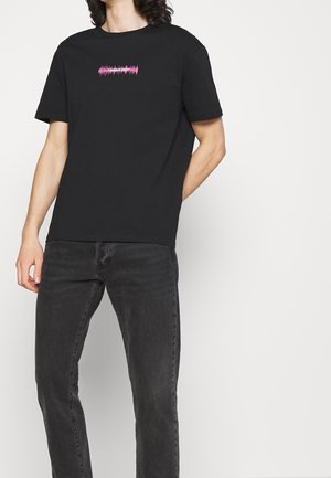 Homme portant un t-shirt noir avec une forme d'onde sonore rose et le texte « shout out », associé à un jean gris foncé, debout contre un mur uni.