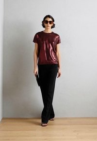 Burgundy metalická tričko v kombinácii s čiernymi nohavicami. Slnečné okuliare a kabelka zvýrazňujú outfit. Hladká textúra, uvoľnený strih, moderná silueta.
