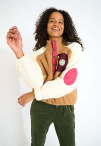 Giacca fluffy marrone e bianca con colletto alto, profilo di bottoni rosa, taschino bordeaux e patch logo rotondo colorato.