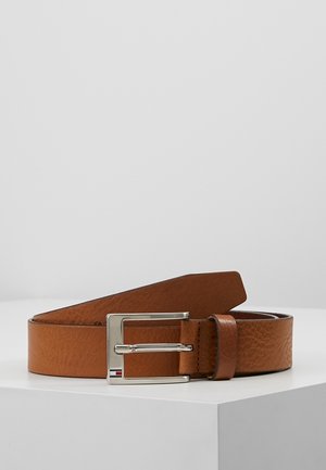 Brązowy skórzany pasek z prostokątną srebrną klamrą i małym czerwono-białym detalem, zwinięty na białej powierzchni.