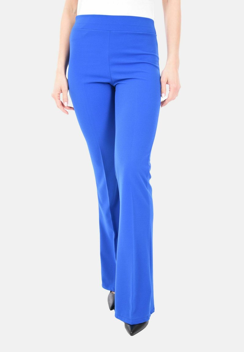 Donna che indossa pantaloni flare fitted blu brillante e scarpe nere a punta, in piedi contro uno sfondo chiaro e neutro.
