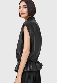Black leather sleeveless top with a peplum hem, featuring a smooth texture and side pockets, shown with a pleated black skirt.