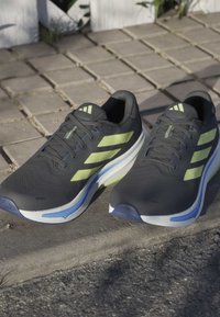 Scarpe sportive nere con righe giallo-verdi, parte superiore in rete testurizzata, suola intermedia bianca spessa e suola esterna blu scuro, con occhielli rinforzati.