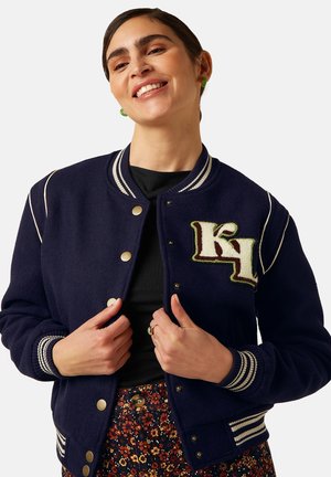 Vrouw die lacht en een donkerblauw varsityjack met gestreepte boorden en een "KL" patch op de borst dichtknoopt, draagt een bloemenbroek en groene oorbellen.