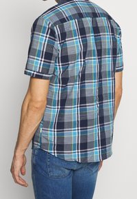 Kortärmad rutig skjorta i nyanser av blått, grönt och grått. Har en knappkrage och en rak fåll med sidorevor.