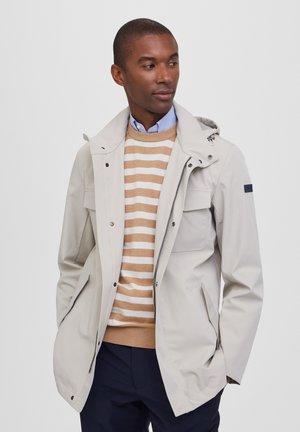 Chaqueta impermeable de color beige claro con capucha, cierre de cremallera y bolsillos laterales, llevada sobre un suéter a rayas en tonos beige y crema, que presenta una camisa con cuello.