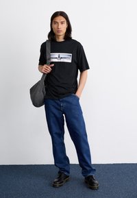 Sort T-shirt med grafik på fronten, denim jeans og sorte sko. Modellen holder en grå ternet skuldertaske. Ensfarvet og afslappet pasform.