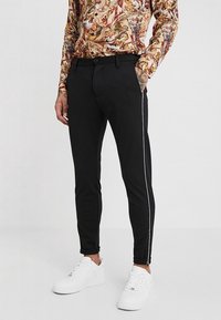 Schwarze Slim-Fit-Hosen mit weißen Seitenstreifen, aus glattem Stoff gefertigt. Mit einem Knopf- und Reißverschluss sowie Fronttaschen ausgestattet. Weiße Sneaker.