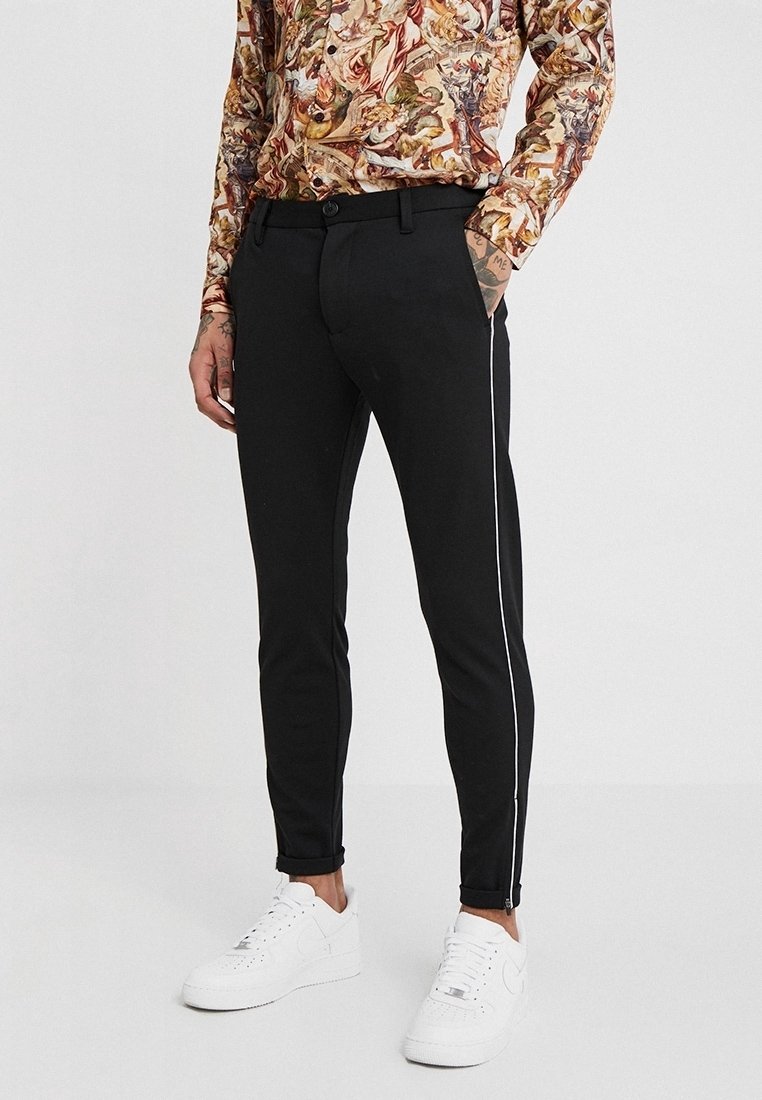 Schwarze Slim-Fit-Hosen mit weißen Seitenstreifen, aus glattem Stoff gefertigt. Mit einem Knopf- und Reißverschluss sowie Fronttaschen ausgestattet. Weiße Sneaker.