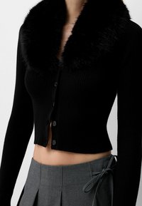 Svart ribbad långärmad kort cardigan med pälskrage, knappad framtill, bärs tillsammans med en högmidjad grå kjol med sidoslående detalj.