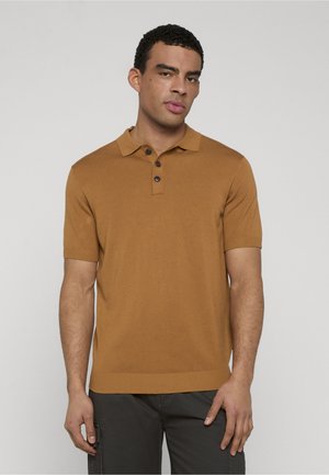 Mann, der ein braunes kurzärmeliges Poloshirt trägt und vor einem einfarbigen hellgrauen Hintergrund steht.